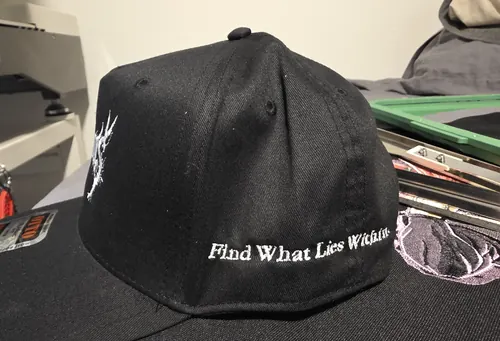Side view of a black Otto cap with the words Find What Lies Within stitched in white on the side