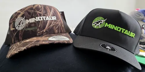 Two caps: a marsh camo trucker with a white Minotaur bull logo and wordmark, and a black performance cap with the logo stitched in neon green