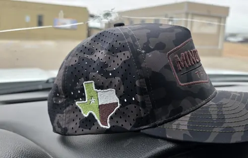 Side view of a dark camo cap with a Texas-shaped flag embroidered on the side panel